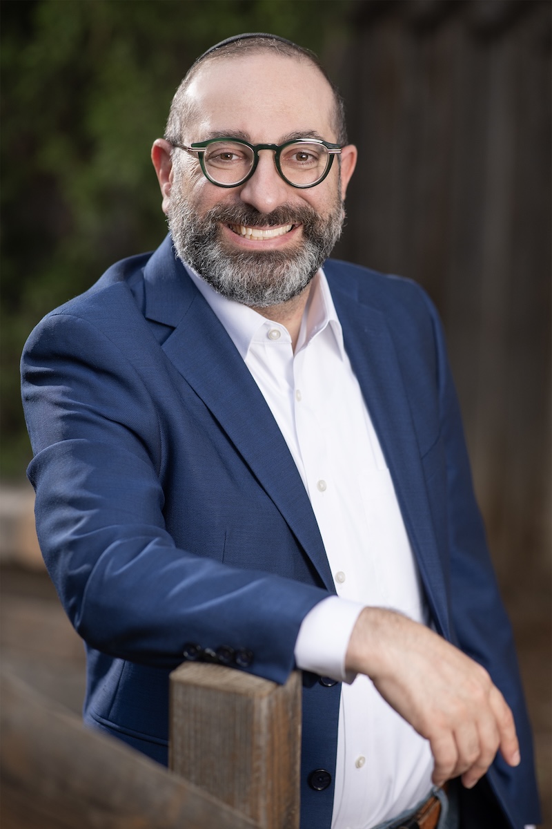 Portrait of Dr. Shimon Blau smiling in a navy suit jacket and white shirt, leaning on a wooden fence outdoors.