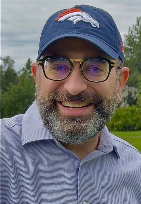 Portrait of Dr. Shimon Blau smiling outdoors, wearing a navy Denver Broncos cap, dark-framed glasses, and a light blue polo shirt.