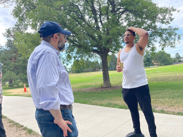El Dr. Shimon Blau, con camisa clara y gorra, conversa con un votante de Denver en un parque en un día soleado.